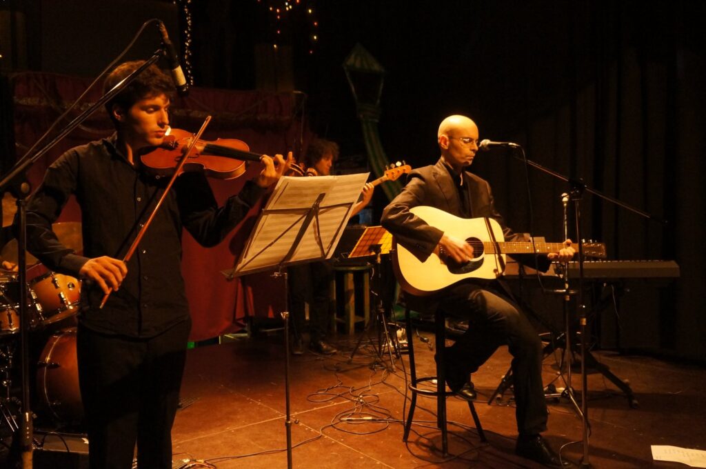 Sur une petite scène éclairée de chaud, un violoniste debout joue devant un pupitre pendant qu'un guitariste assis chante au micro ; batterie, contrebasse et claviers occupent l'arrière-plan.