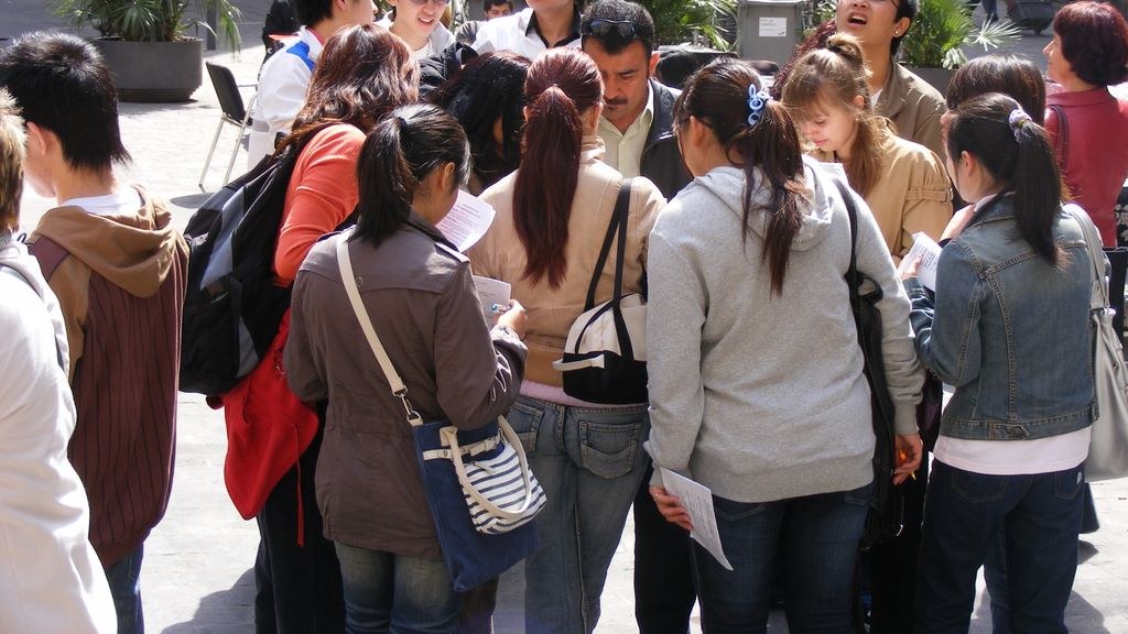 Un groupe d’étudiants adultes de l’Escola Oficial d’Idiomes, vus de dos, réunis sur une place de Barcelone, feuilles d’exercice à la main, pendant une sortie de classe dans le quartier de Sant Pere.