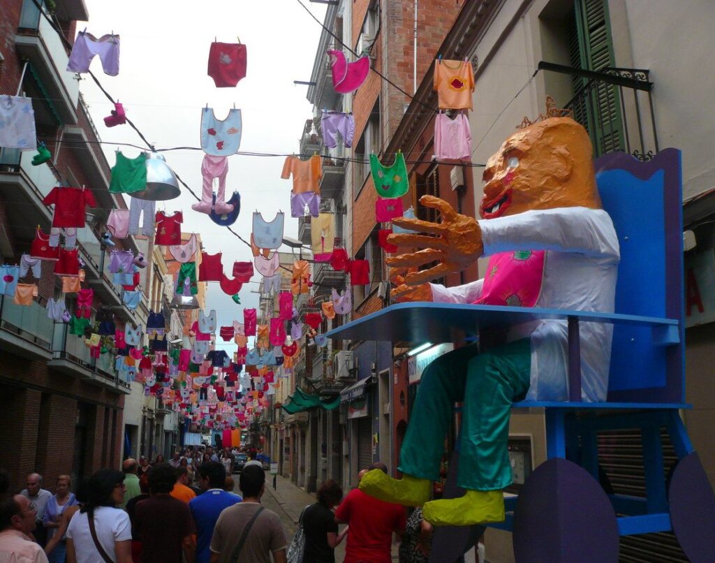Une rue de Sants décorée pour la festa major : guirlandes de petits vêtements colorés tendues au-dessus de la chaussée et immense personnage en papier mâché assis sur une chaise bleue, au milieu de la foule.