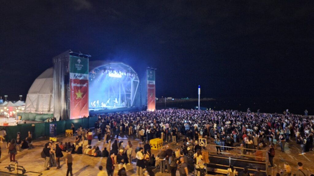 Une scène de concert éclairée en bleu sur la platja del Bogatell, de nuit, pendant la Mercè, avec une foule dense devant elle et la mer à droite.