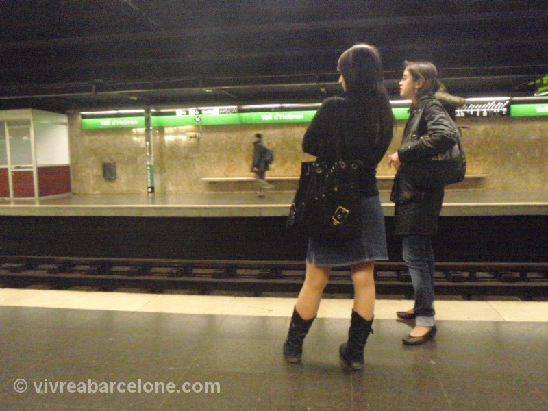 Deux voyageuses attendent sur un quai du métro de Barcelone, devant les panneaux verts de direction de la ligne, un troisième voyageur assis sur le banc du quai d'en face.