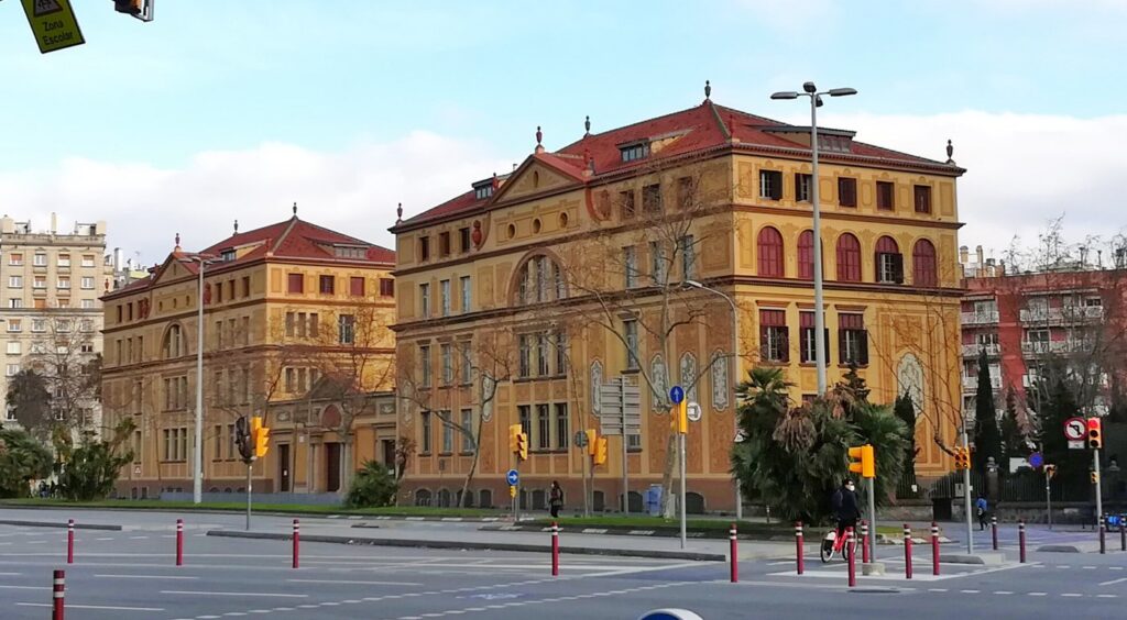 La façade ocre et les toits rouges du Grup Escolar Ramon Llull, avenue Diagonal à Barcelone : une des grandes écoles publiques de la ville, sous un ciel bleu, avec un panneau « Zona Escolar » au premier plan.