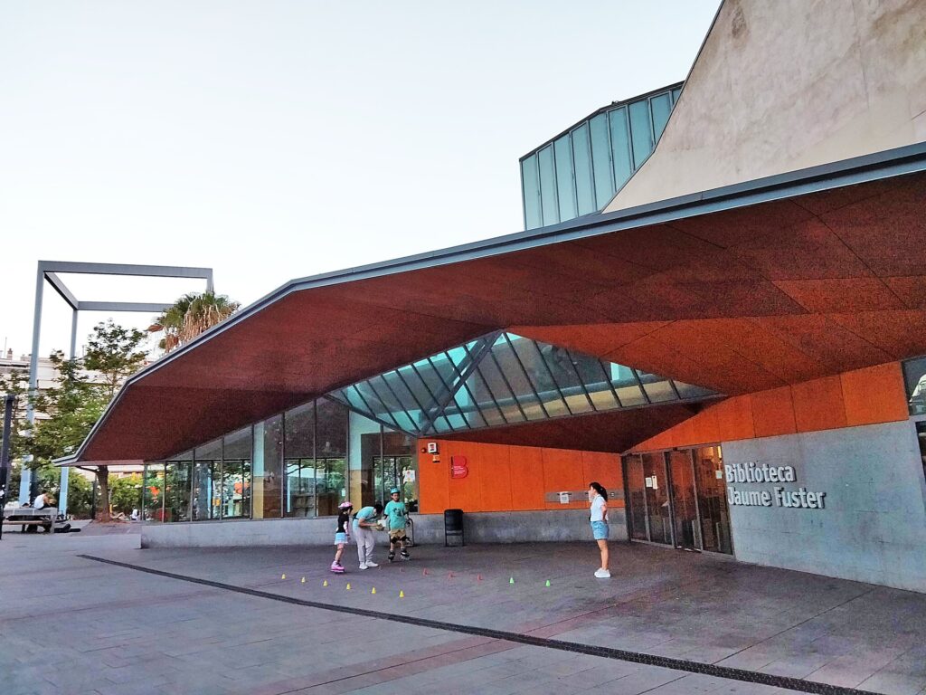 L'entrée de la Biblioteca Jaume Fuster, place Lesseps à Barcelone : un grand auvent de pierre rouge au-dessus d'une façade vitrée, avec le nom de la bibliothèque gravé sur le mur, et quelques passants devant l'esplanade.