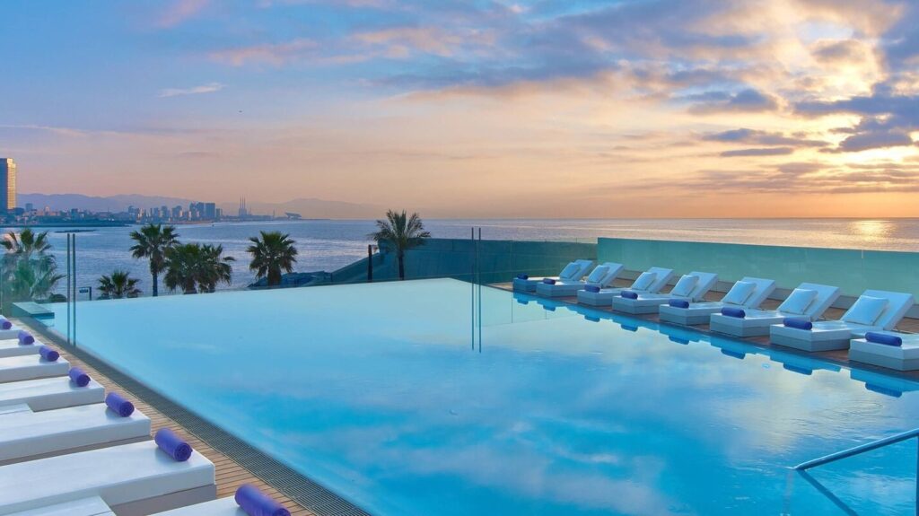 Piscine à débordement d'un hôtel de bord de mer à Barcelone au coucher du soleil, avec les tours du Port Olímpic au loin